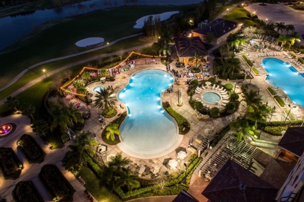 An aerial view of three pools at night, surrounded by a lit patio and strings of bistro lighting. 