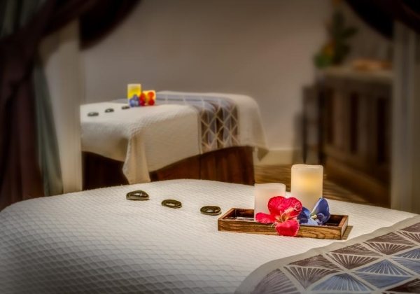 Two massage tables adorned with a tray with a candle, flowers, and hot stones in a serene spa setting. 