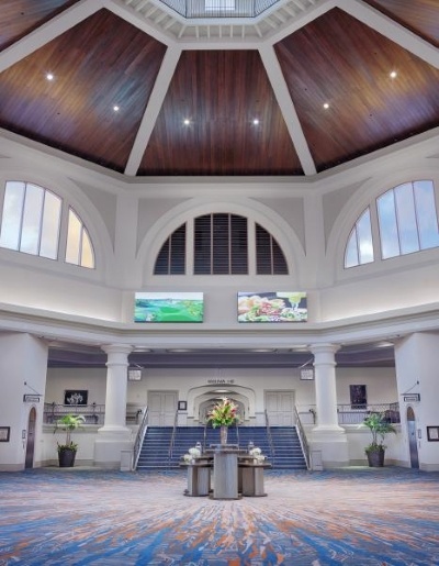  A spacious reception area with blue carpet set with a display of floral arrangements in front of a small flight of stairs. Arched windows two stories above lead the eye to a rich, dark wood rotunda ceiling. 