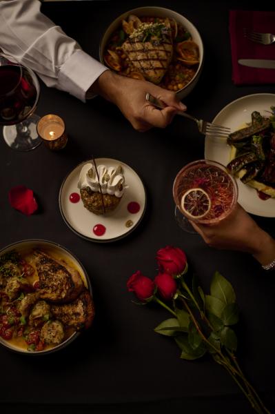 An aerial view of a dimly lit table with a hand holding a fork, another holding a cocktail with an orange slice, a plated dessert, roses, and other plates of food. 