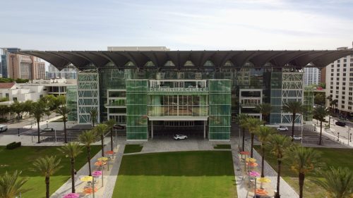 An image showing the Dr. Phillips Center (a large building with a flat roof, full-length glass windows, and a front lawn lined with palm trees and pink, yellow, and orange umbrellas. Catch these amazing events at the Dr. Phillips center this January while you stay in Orlando.