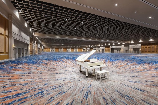 A large ballroom with blue and rust-patterned carpet that is completely empty except for a white baby grand piano. 