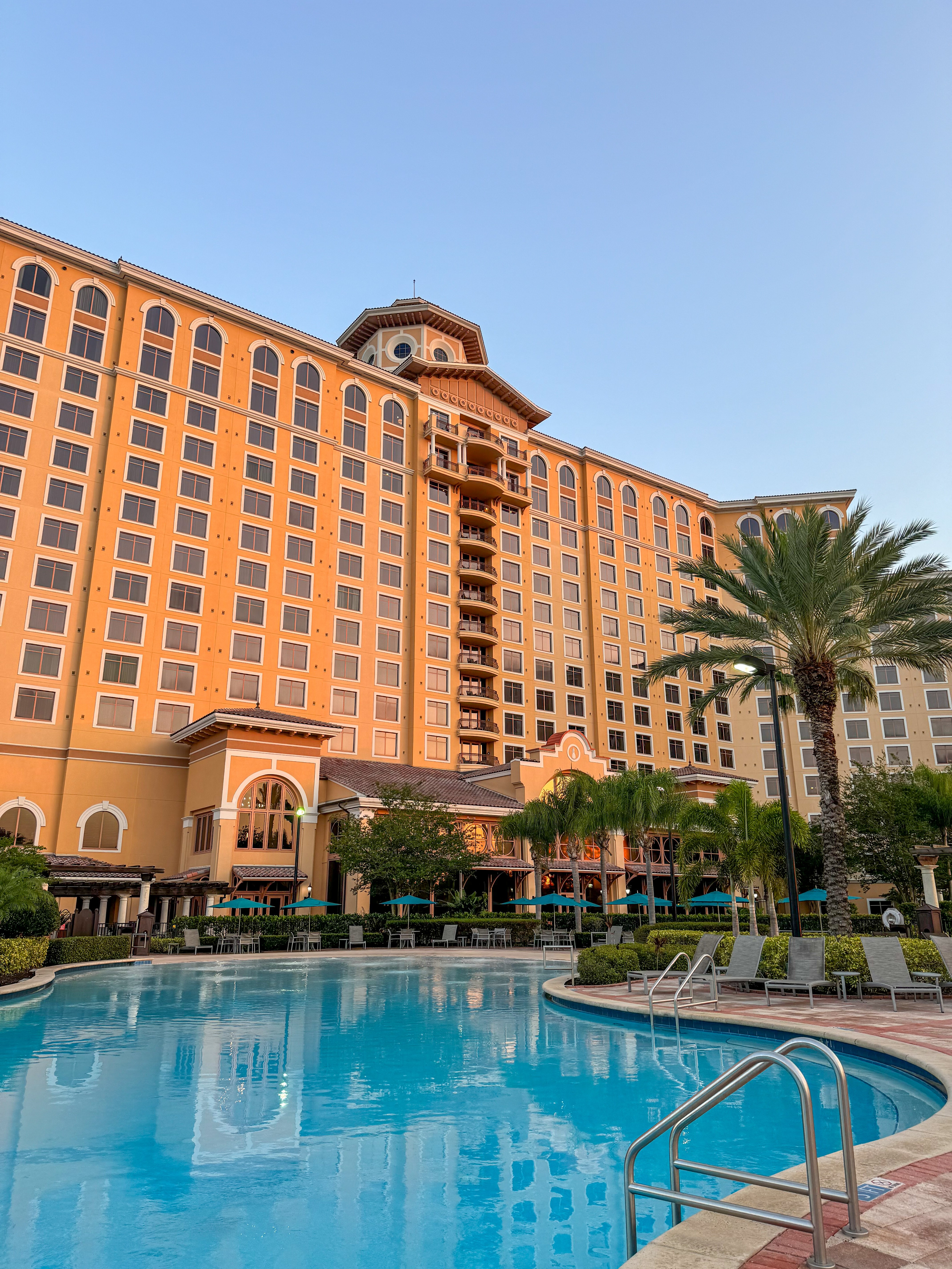 A hotel with facade with more than 100 windows, a pool in front with palm trees and beach chairs surrounding the pool. After a night of entertainment at Dr. Phillips Center in Orlando, enjoy a relaxing and luxurious stay at Rosen Shingle Creek.