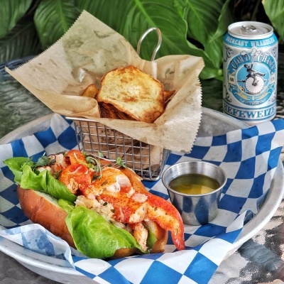 A lobster roll with lettuce, a cup of butter, and a basket of potato chips sit on a sheet of blue checkered tissue paper beside a can of beer. The Nauti Lobstah serves some of the best seafood in Orlando.