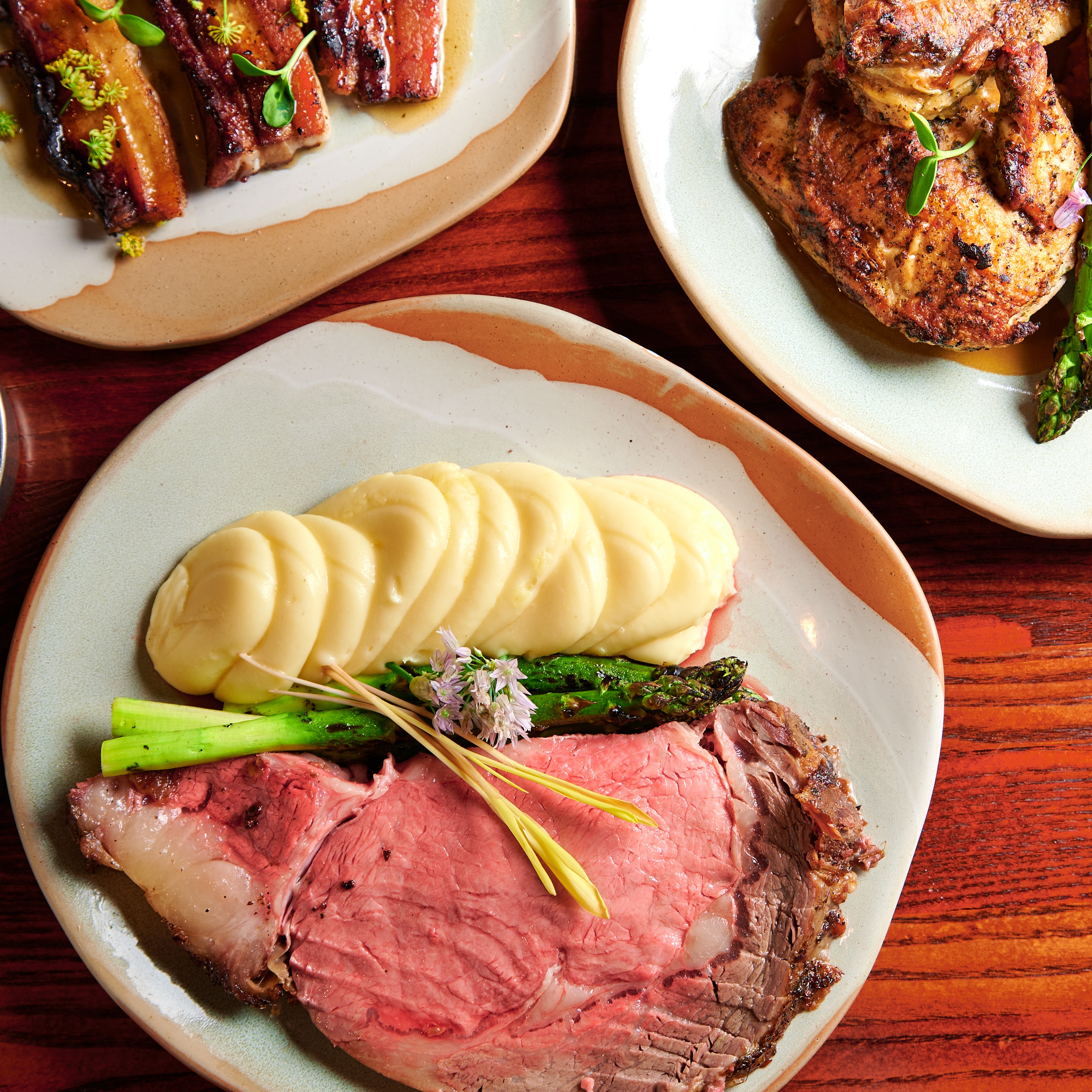 A white dish with prime rib, asparagus, and mashed potatoes served in an elegant manner, surrounded by two other cut off dishes of food. 