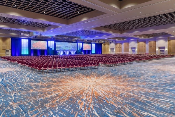 A large open ballroom with a blue and rust patterned carpet set with a large, blue-lit stage and rows of hundreds of red cushioned chairs. 