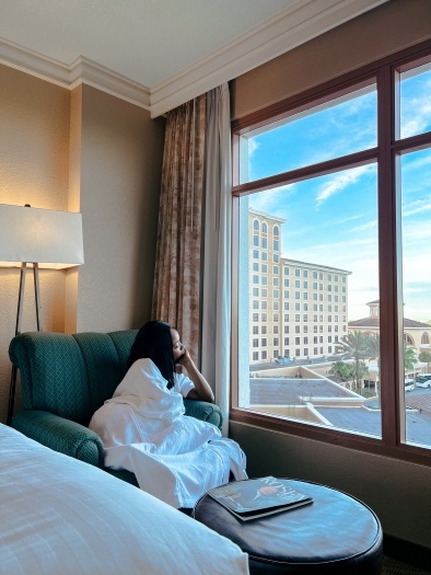 A woman sits in an armchair staring out the window with a large hotel in the distance.