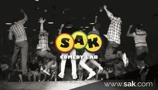 A black and white image of six performers jumping onstage with their backs to the audience, overlaid with the SAK Comedy Lab logo. 